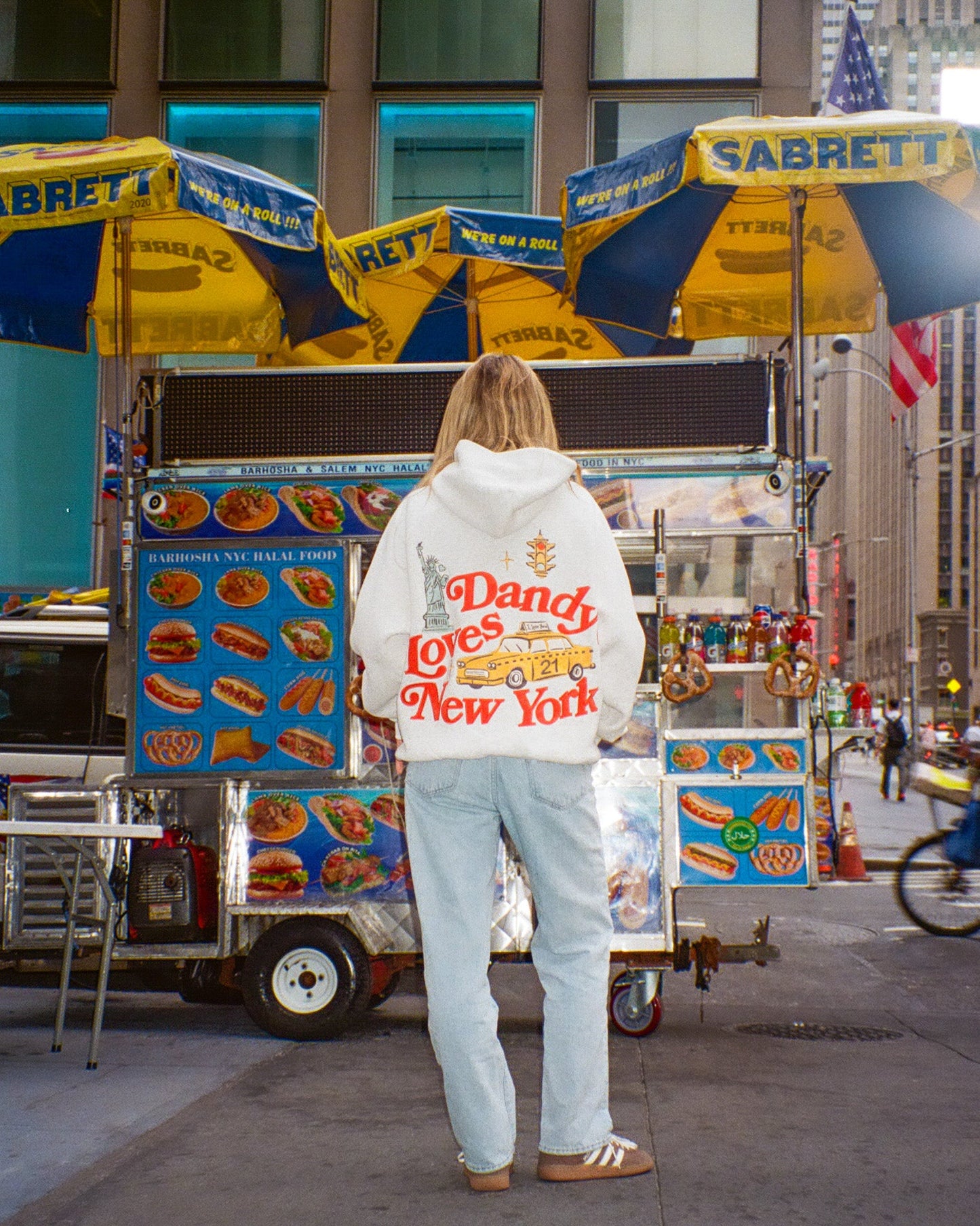 Person wearing a hoodie with 'Dandy Loves New York' text in front of a Sabrett hot dog cart.